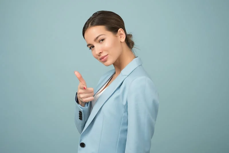 A woman in a blue suit is pointing to her face.