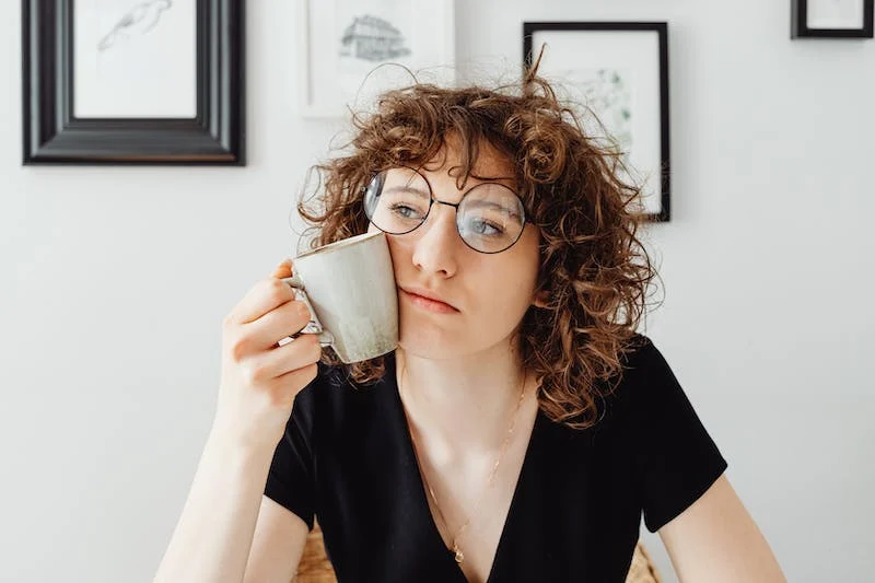 A woman with curly hair and glasses holding a cup.