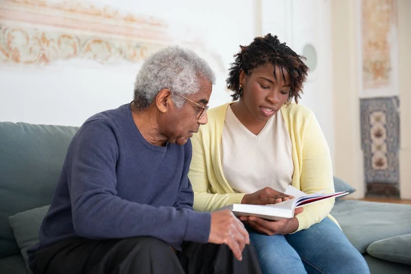 A man and woman sitting on the couch looking at an open book.